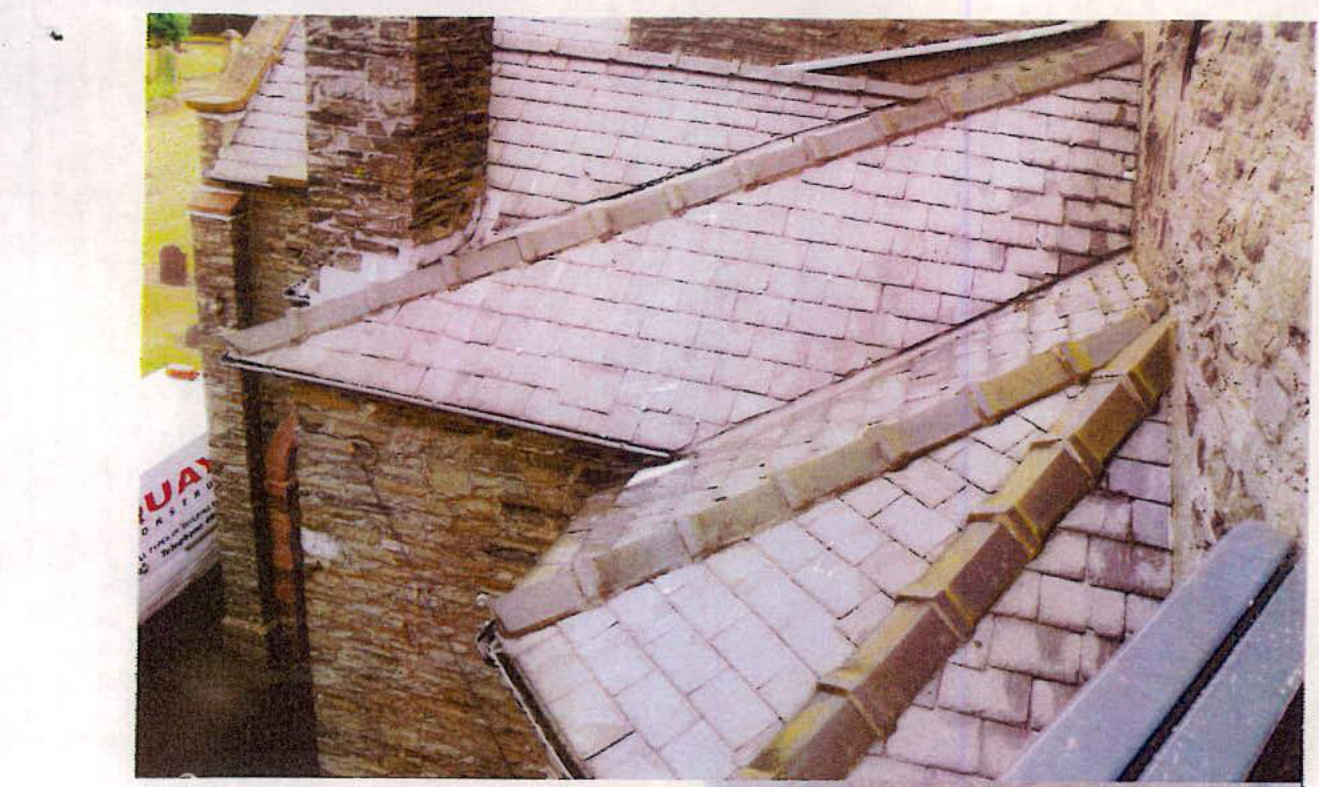 A close-up photograph showing a slate roof with brick chimneys and ridges, likely documenting the existing state before chimney removal.