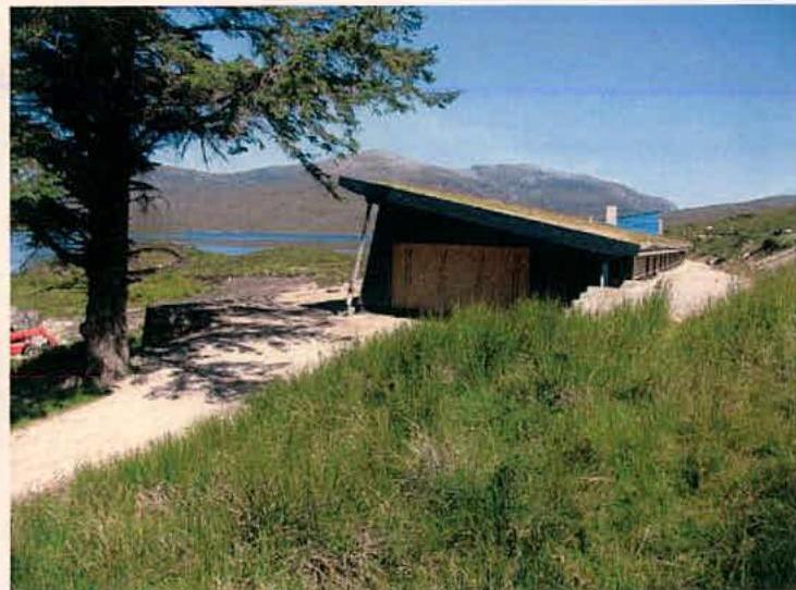 A photograph showing a single-story building with a sloping roof, likely a visitor centre, situated in a grassy, rural landscape near water and hills.