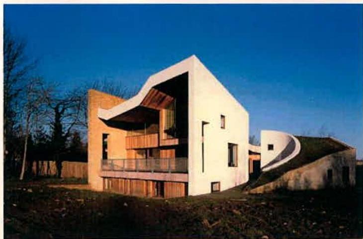 A photograph or artist impression of a modern building featuring a white angular facade, wooden cladding, and a grassy green roof section.