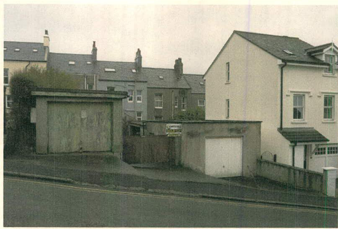 A street-level photograph showing existing residential properties and garages, including a detached garage on the left and a white house on the right, with terraced housing in the background.