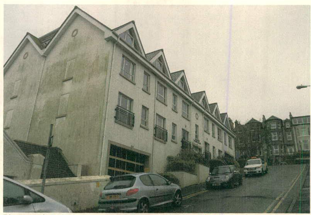 A photograph showing a large, white, multi-story residential building with a series of gables and balconies, situated on a street with parked cars.
