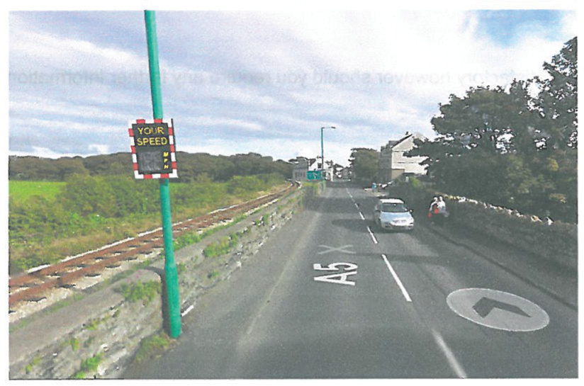 A photograph showing a street scene on the A5 road in Castletown, featuring a speed camera sign, a parallel railway line, and nearby buildings.