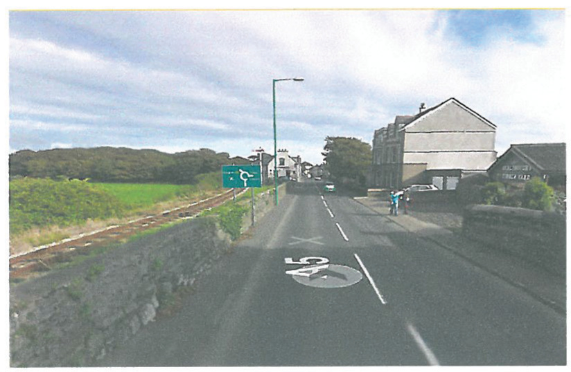 A street-level photograph showing a road with a roundabout sign, speed limit markings, and buildings on the right side.