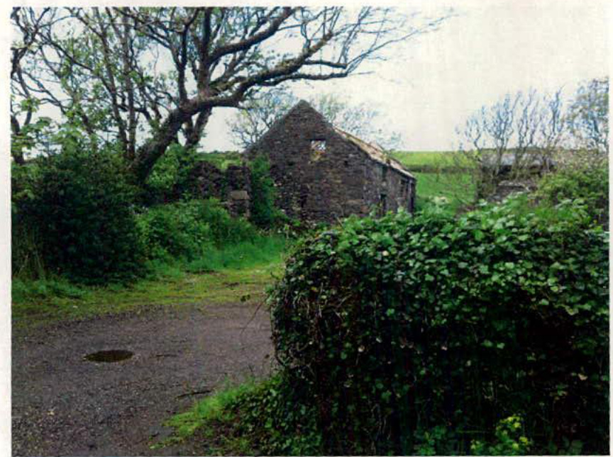 A photograph depicting a dilapidated stone barn structure situated in a rural, overgrown landscape with a gravel path in the foreground.