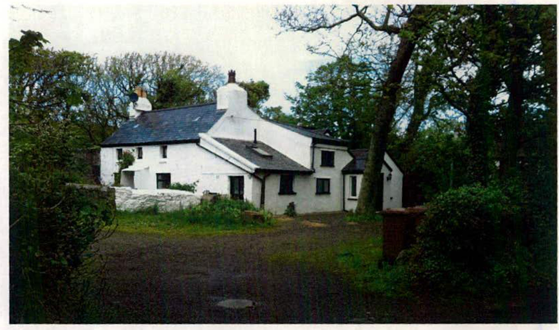 A photograph showing a white, single-story building with a slate roof situated in a rural setting with surrounding trees and a gravel driveway.