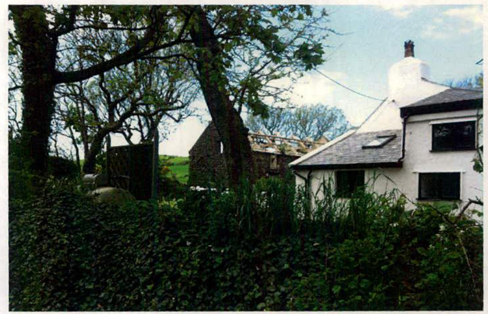 A photograph showing a white detached house on the right and a dilapidated stone barn structure in the background amidst trees.