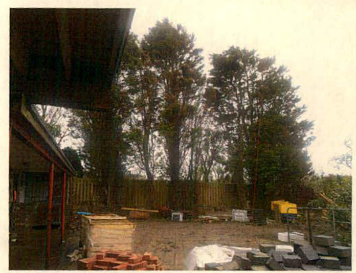 A photograph showing a garden area under construction with building materials like bricks and slabs in the foreground, a wooden fence, and tall trees in the background.