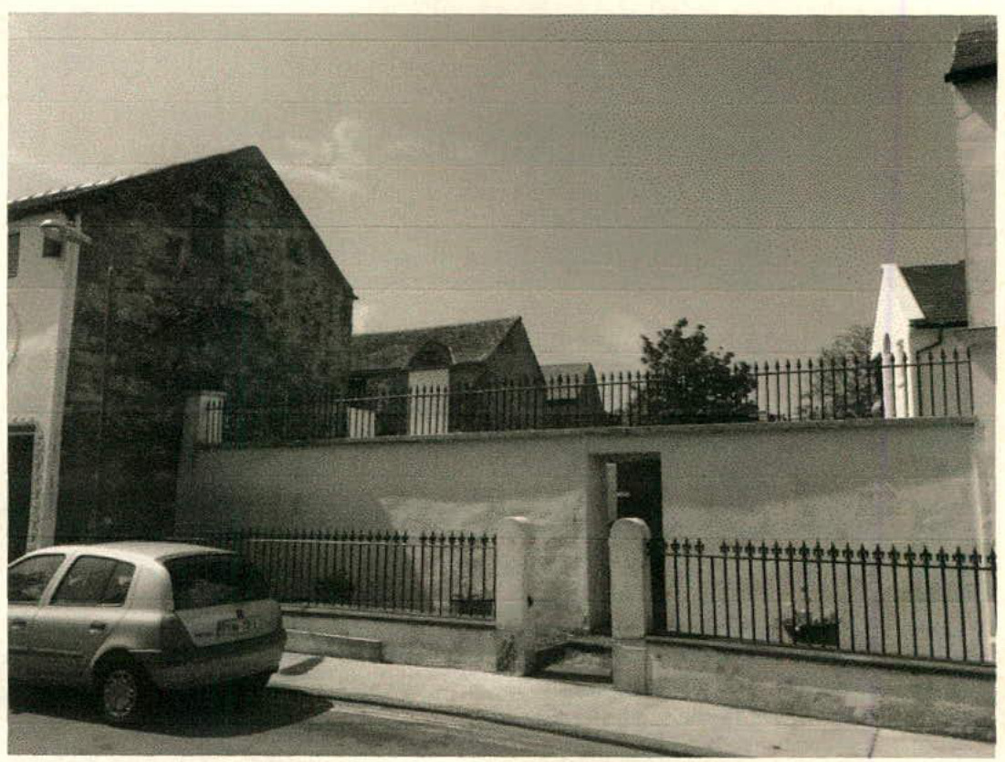 A black and white photograph showing a street view of a stone building, a boundary wall with a gate, and a parked car.