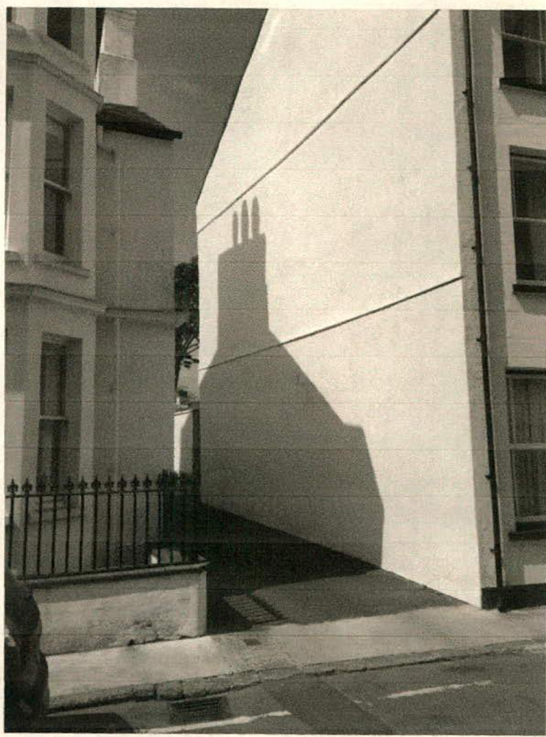A black and white photograph showing a narrow gap or alleyway between existing buildings, featuring a prominent shadow cast on a white wall.