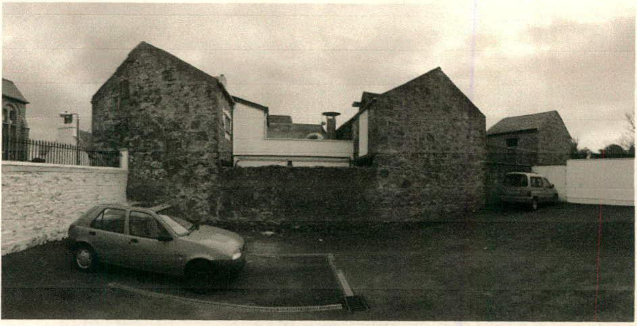 A black and white photograph showing the exterior of a stone building with a white extension section, with cars parked in the foreground.