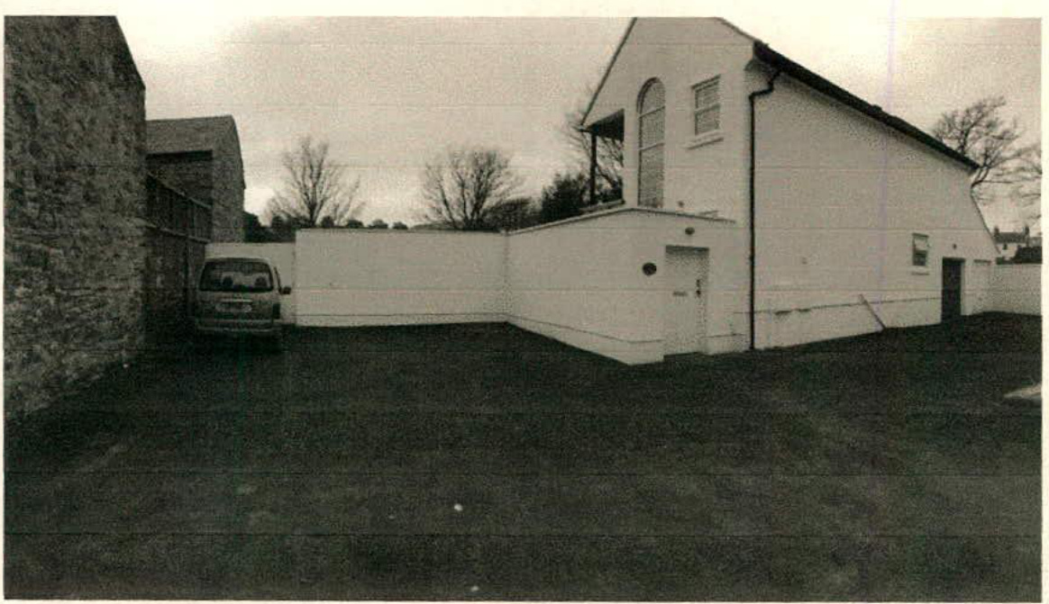 A black and white photograph showing a white two-story building with a pitched roof and large arched window, situated next to a white boundary wall and a stone wall with a parked van.