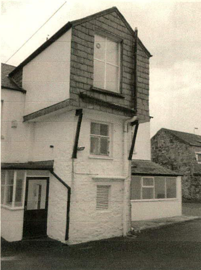 A black and white photograph of a white-washed stone building featuring a prominent two-storey extension with a slate roof and large window.