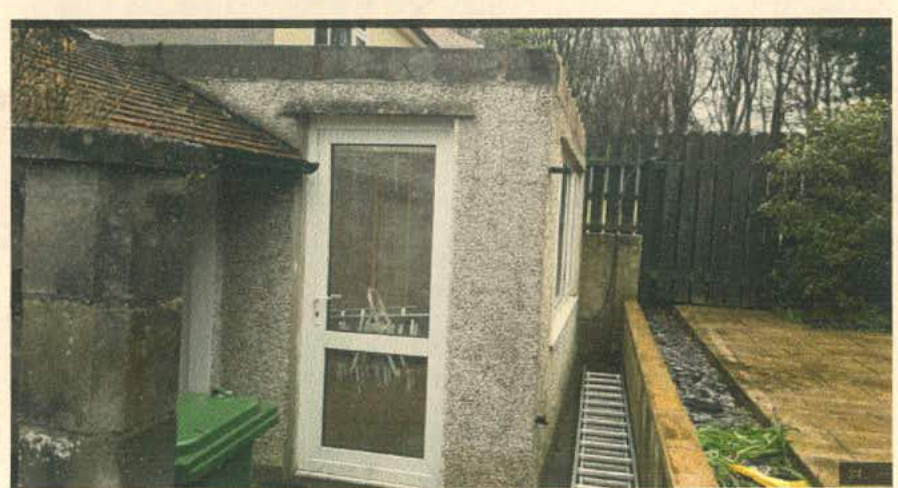 A photograph showing the side elevation of a single-story building with pebbledash rendering, a white door, and a flat roof area.
