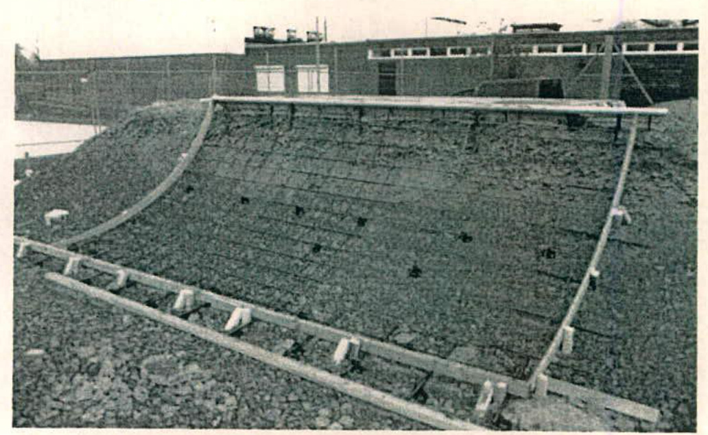 A black and white photograph depicting a large concrete skate ramp or bowl structure with wooden formwork along the edges, situated on a gravel surface.