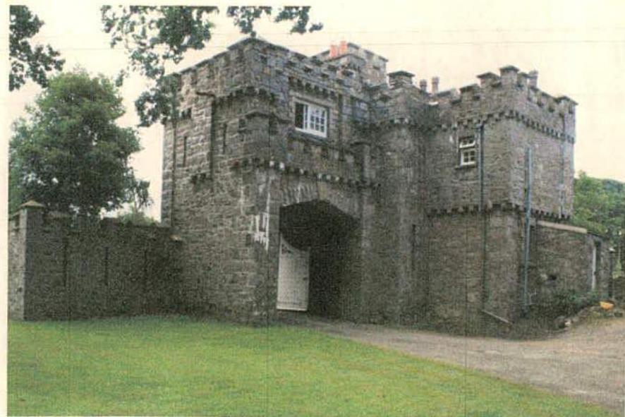 A photograph of a historic stone gatehouse featuring crenellated towers and a central arched gateway with a white gate.
