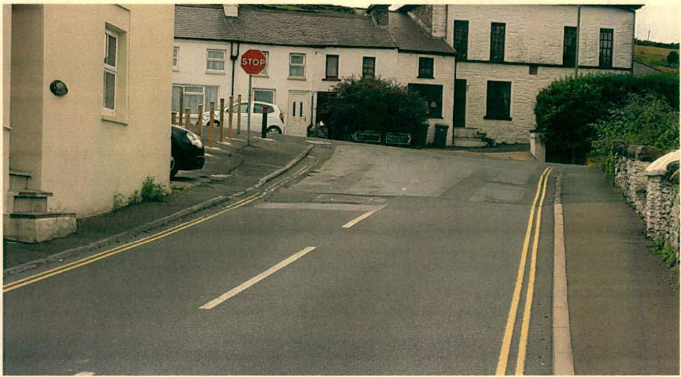A street-level photograph showing a road junction with a stop sign, double yellow lines, and white stone buildings typical of a rural village setting.