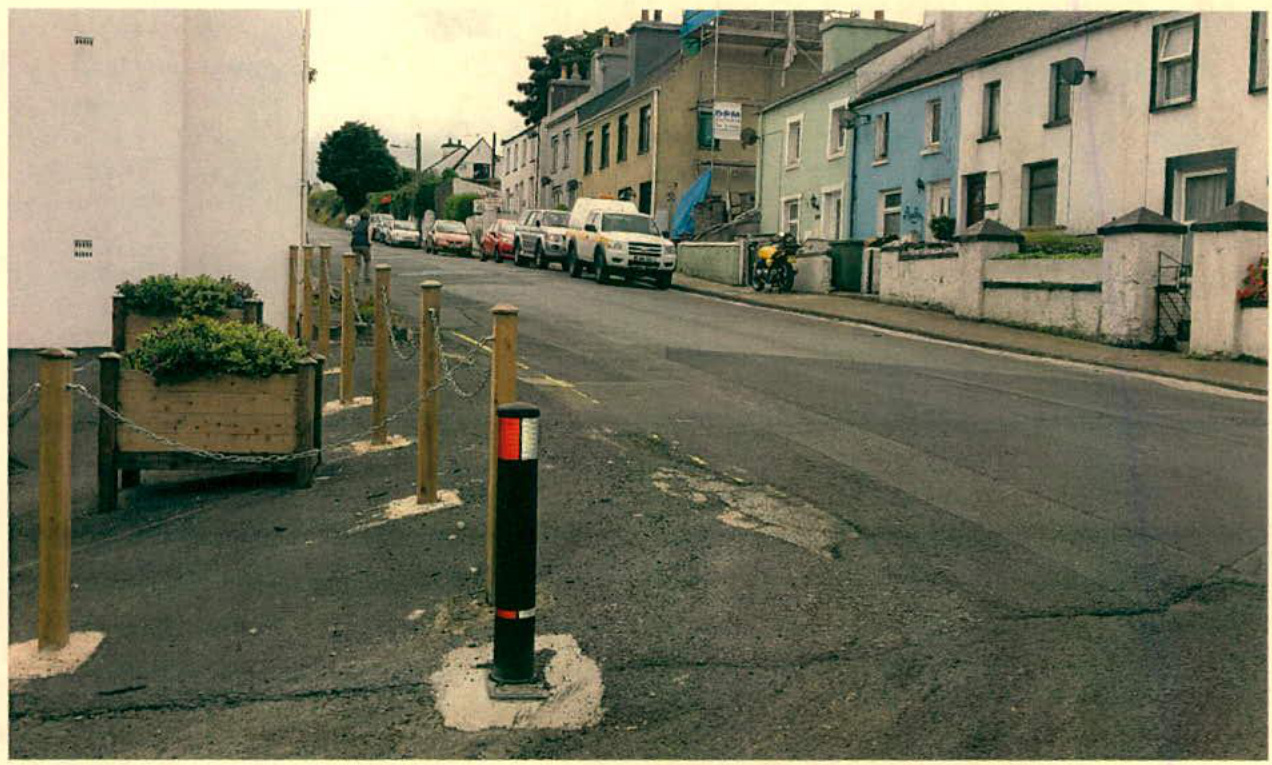 A street scene photograph showing a paved road with parked vehicles, a row of terraced buildings, and foreground landscaping features including wooden planters and bollards.