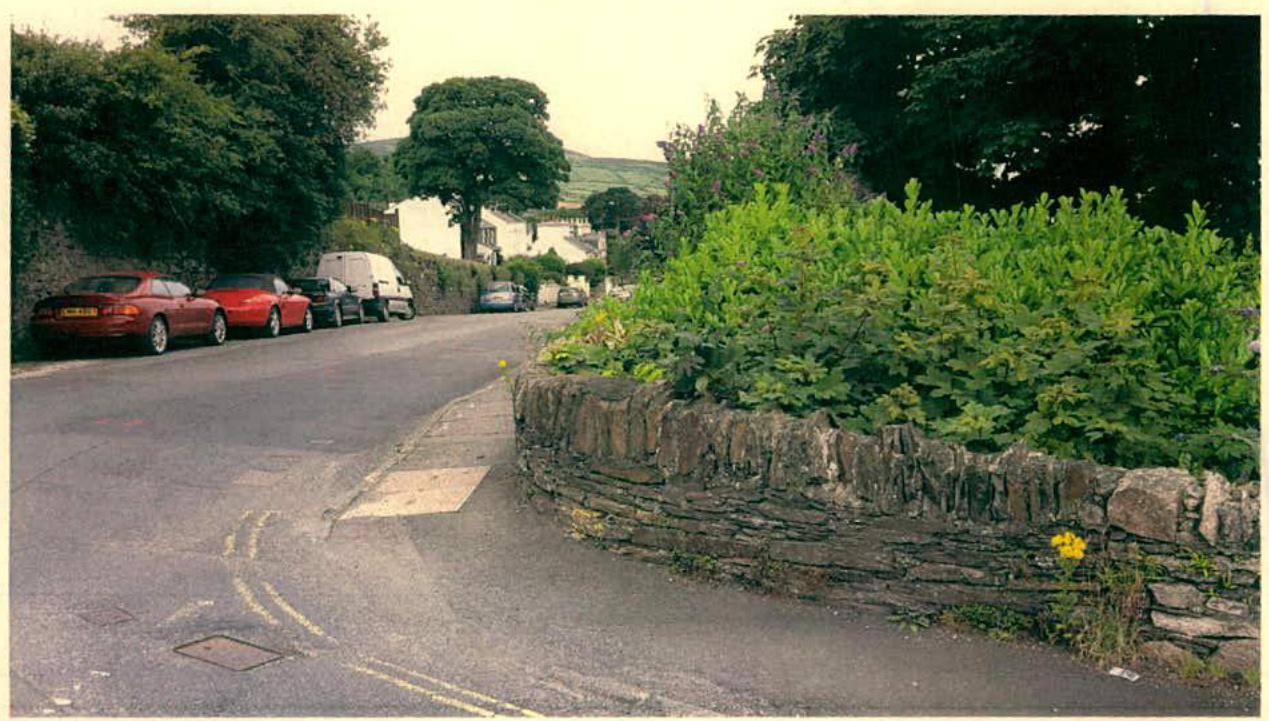A photograph showing a rural road scene with parked cars on the left and a stone wall with vegetation on the right.