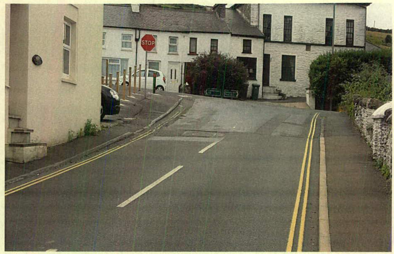 A street-level photograph showing a road with double yellow lines, a stop sign, and white stone buildings on either side, likely depicting the site of the proposed highway improvements.