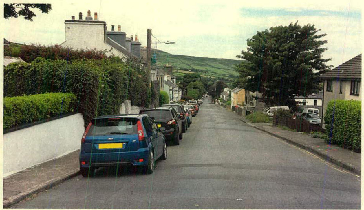 A street-level photograph showing a row of parked cars along a residential road leading uphill towards a green hill.