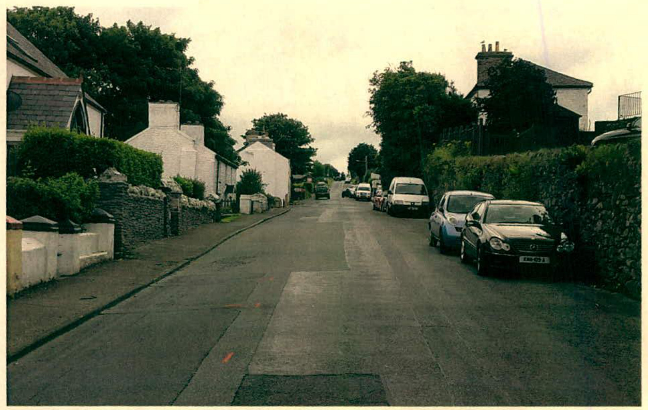 A street-level photograph showing a road with cars parked along the right side, flanked by stone walls and hedges.