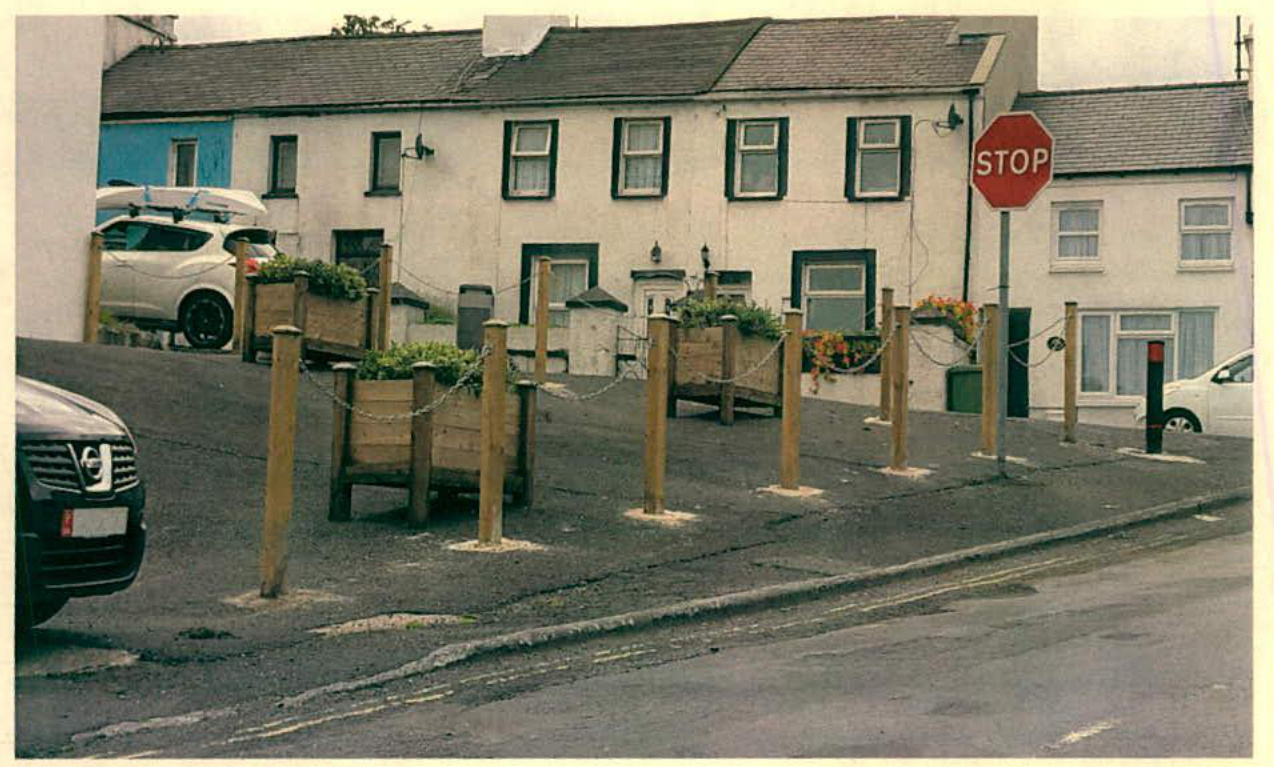 A street-level photograph showing a paved area with wooden bollards and planters in front of a row of white terraced houses.