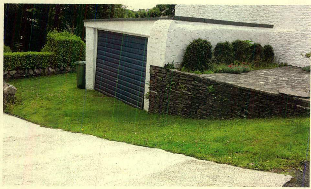 A photograph showing a white garage structure with a dark roller door, adjacent to a stone retaining wall and grassy verge.