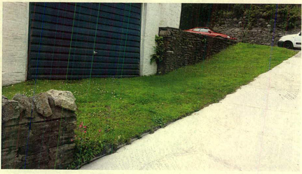A photograph showing the existing vehicular access and driveway leading to a garage with a large dark door. The image captures the stone boundary walls, grassy slope, and parked cars relevant to the proposed highway i...