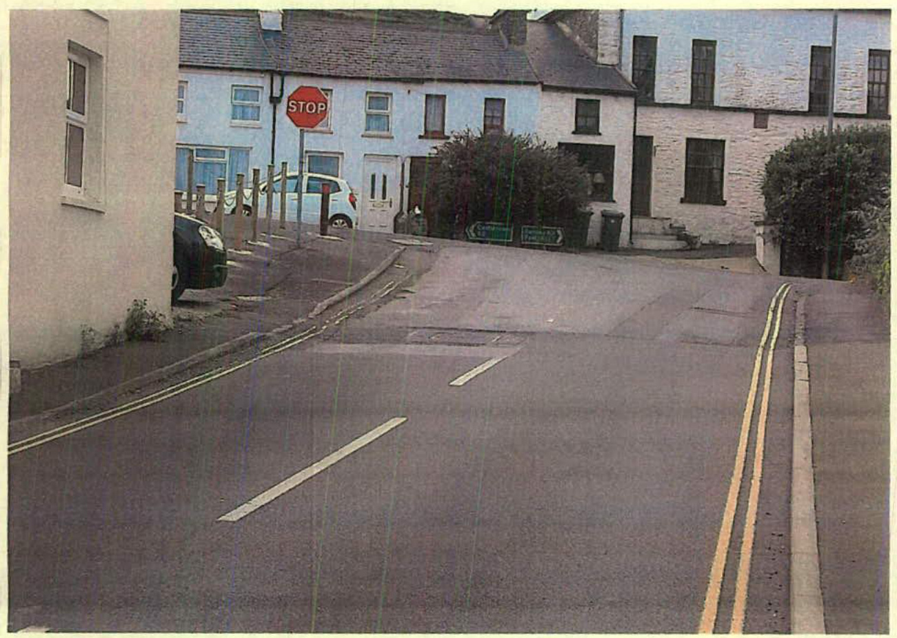A street-level photograph showing a road junction with a stop sign, terraced houses, and parked vehicles.
