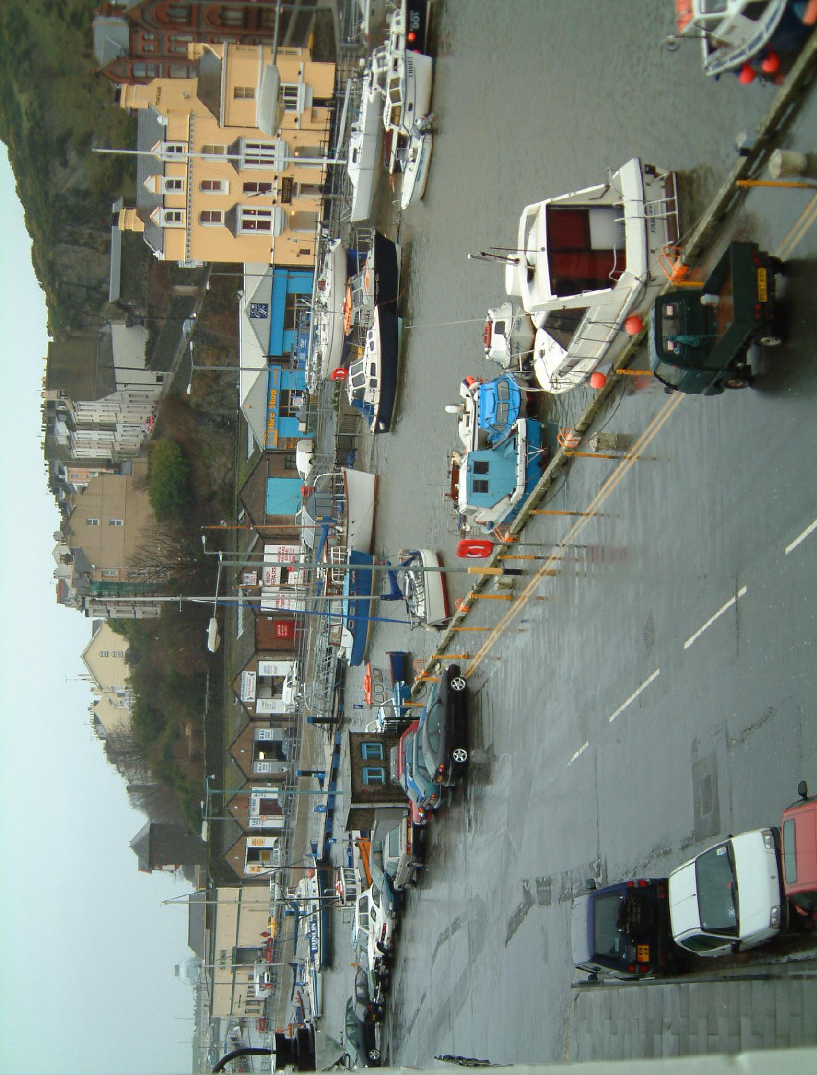 A photograph showing a harbor scene with boats docked along a quay, parked cars, and buildings on the adjacent hillside.