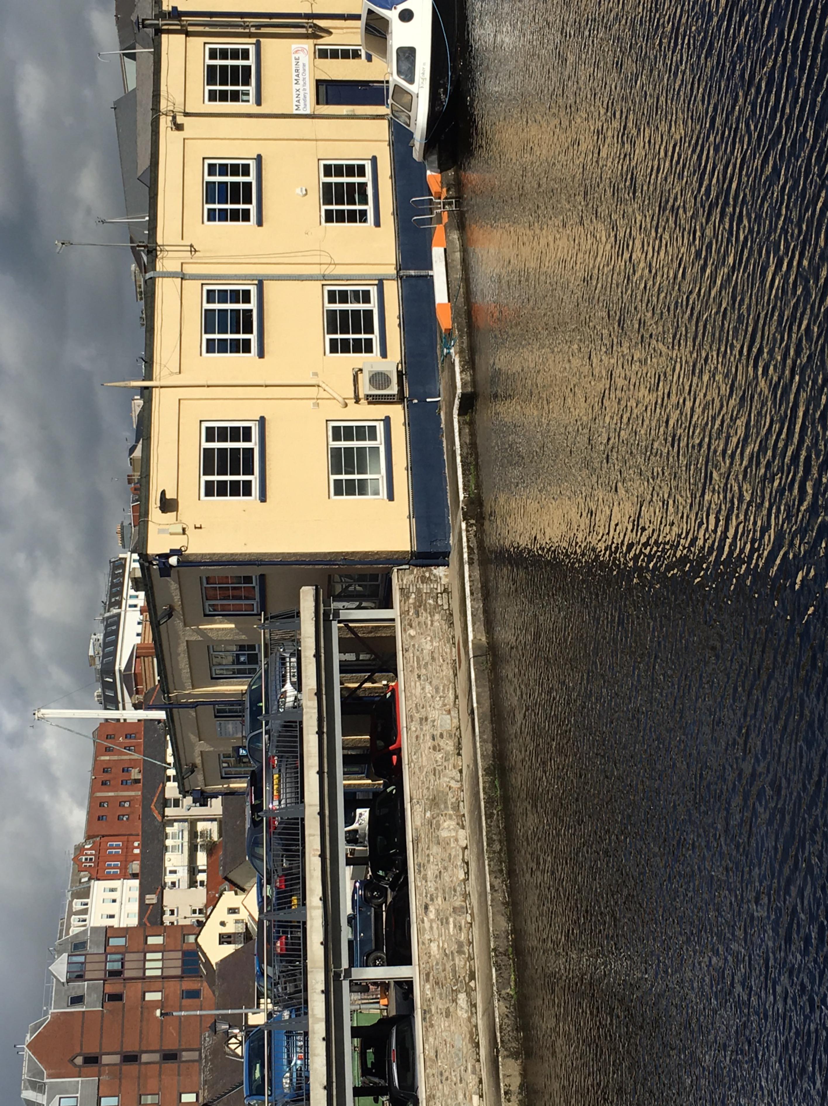 A rotated photograph showing a yellow multi-story building adjacent to a boat and water, with a parking area and other buildings in the background.