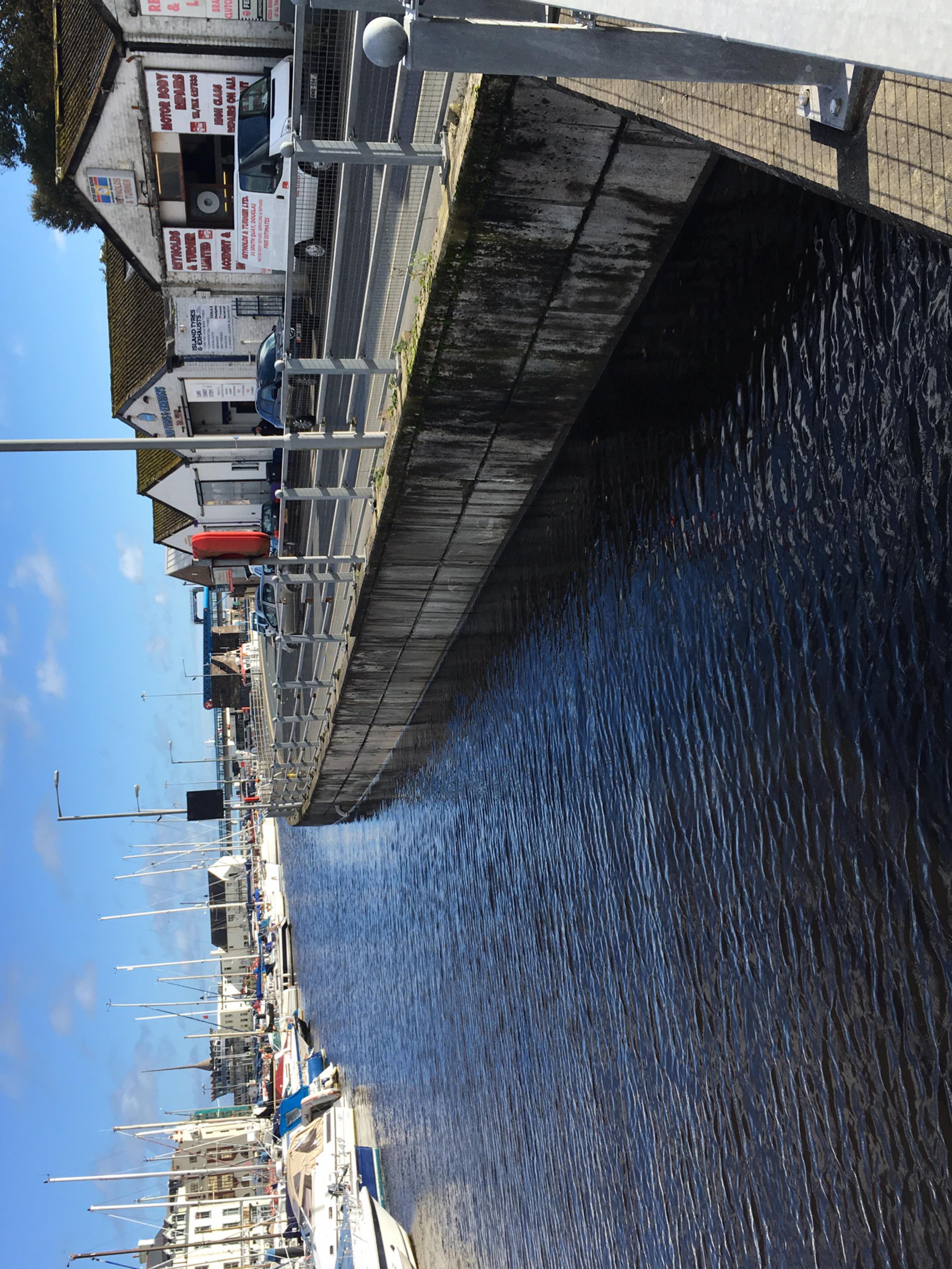 A photograph showing a harbor quay with commercial buildings, parked cars, and moored boats along the waterfront.