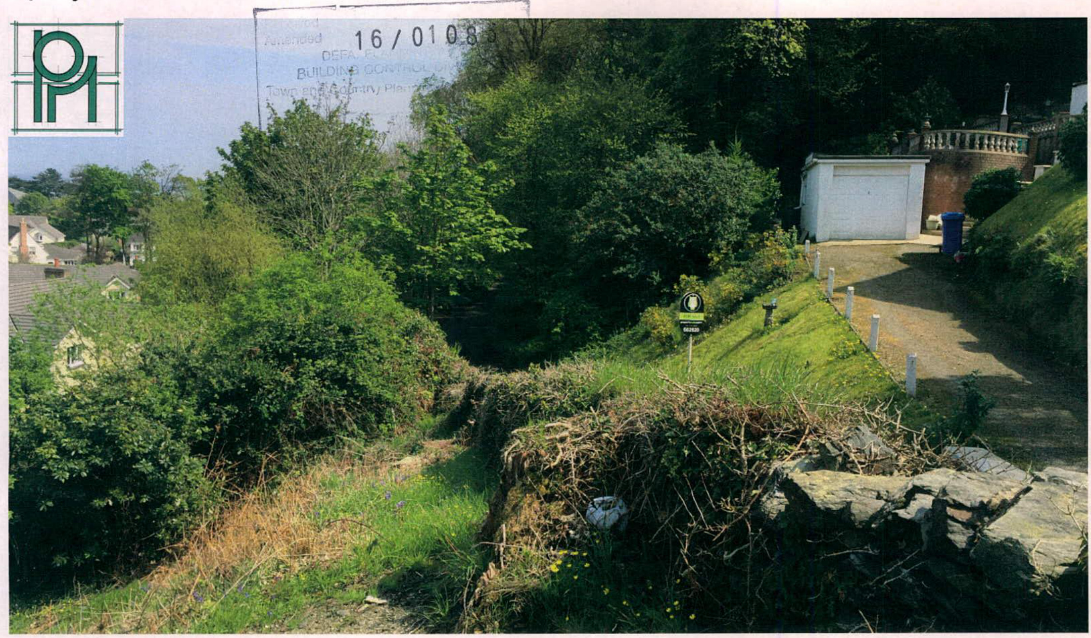 A photograph of a sloping, vegetated site featuring a stone wall in the foreground and a driveway leading to a white garage on the right.