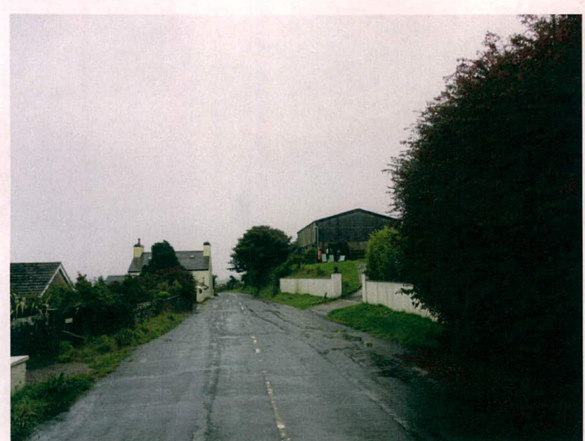 A photograph showing a rural road scene with existing houses on the left and a large agricultural building on a hill to the right.