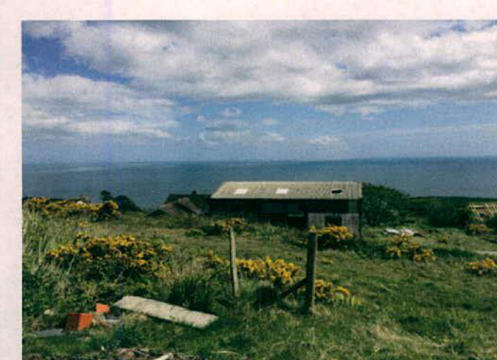 A photograph showing a grassy coastal site with gorse bushes in the foreground and a single-story building overlooking the sea.