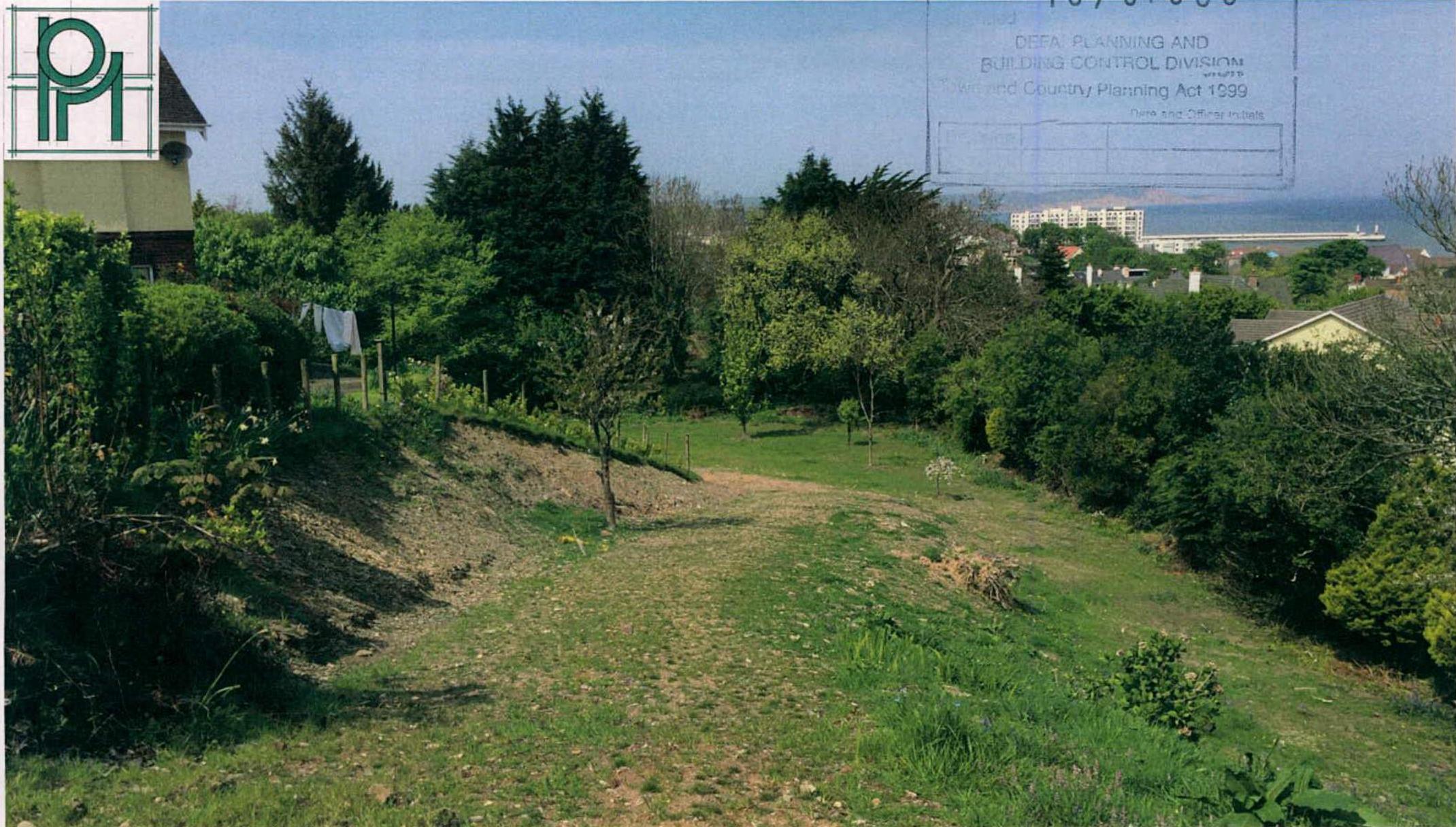 A photograph of a grassy, sloping plot of land with trees and vegetation, overlooking a coastal area with buildings in the distance.