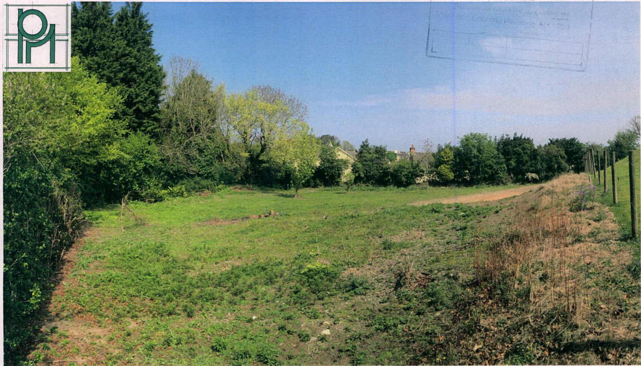 A photograph of a grassy plot of land bordered by trees and a fence, showing the current state of the site.