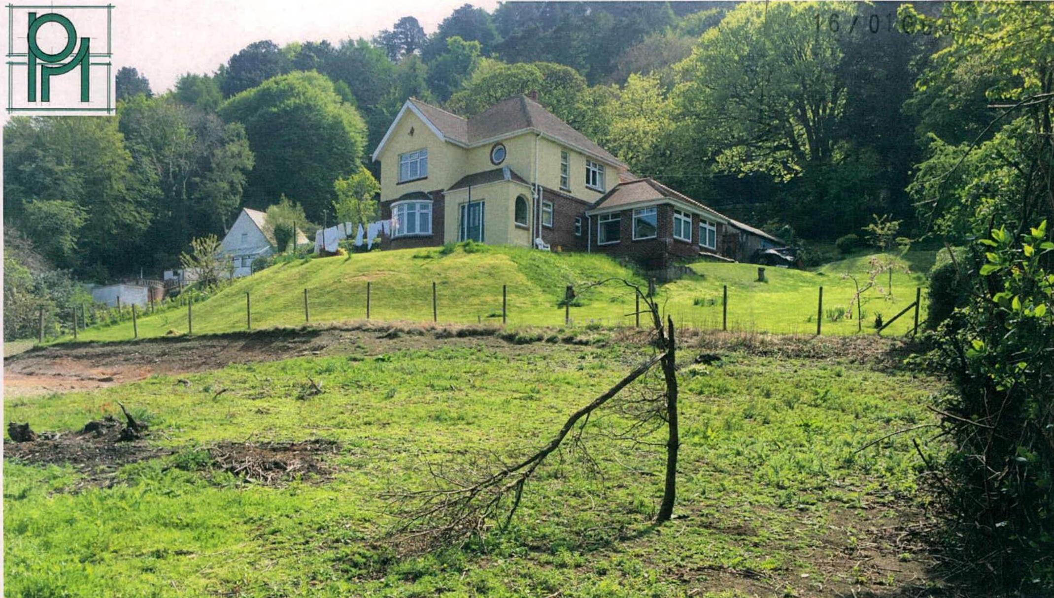 A photograph showing a large detached house situated on a grassy slope with dense trees in the background and a fence line in the foreground.