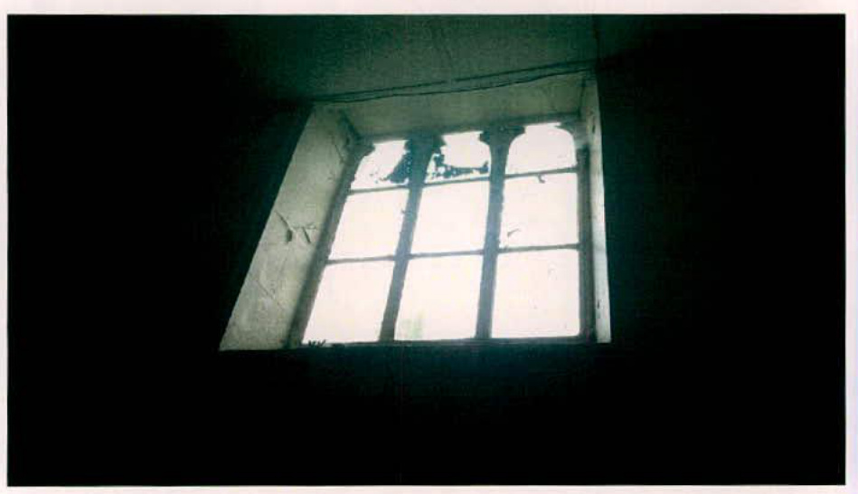 A low-light interior photograph showing a tall, multi-paned window with a stone surround looking out into bright daylight.