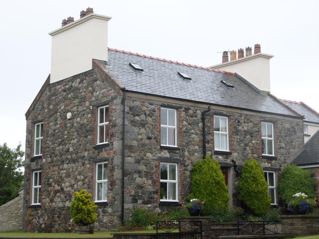 A photograph of a two-story stone house with a slate roof, white window frames, and chimneys, situated in a rural setting.