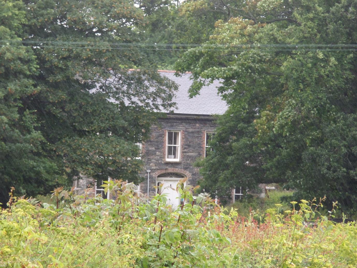 A photograph showing a stone house partially hidden by mature trees and overgrown vegetation in the foreground.