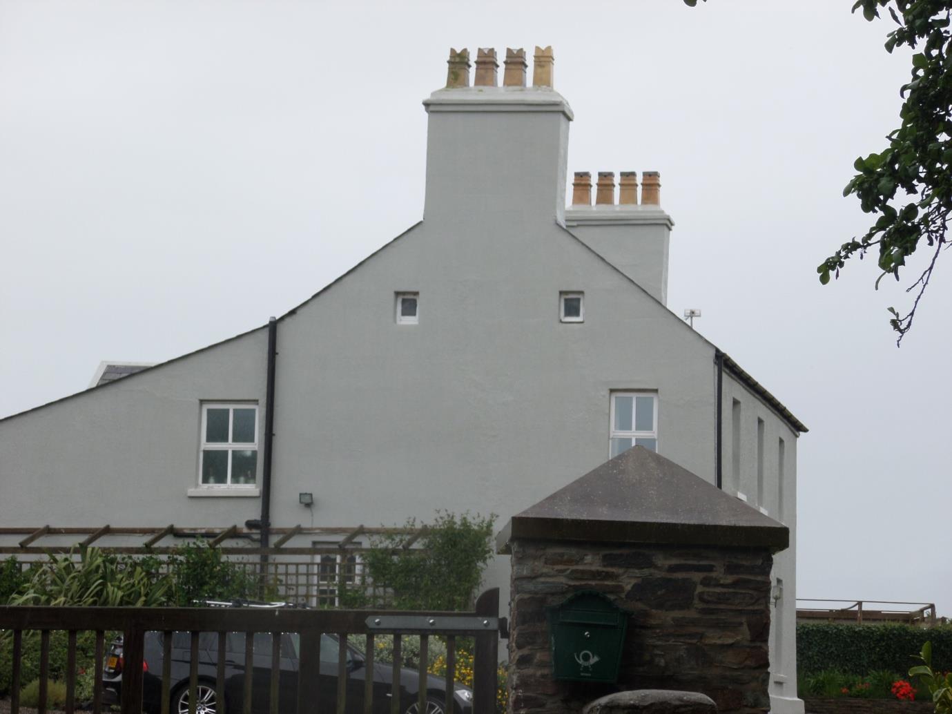 A photograph of a white, two-story detached house with prominent chimneys and small attic windows, viewed from the garden with a stone wall and fence in the foreground.