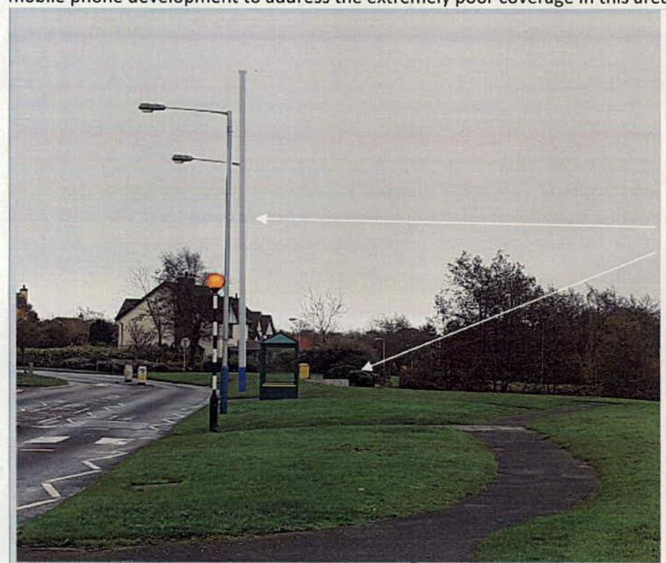 A street-level photograph showing a tall white pole (the proposed antenna mast) standing next to an existing street lamp and a bus shelter on a grassy roadside verge.