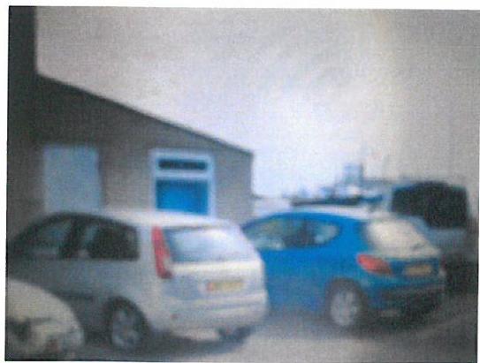 A grainy, low-resolution photograph showing the exterior of a building with a blue door and cars parked in the foreground.