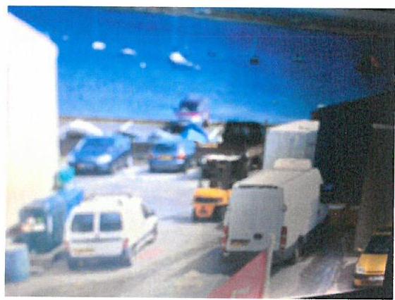 A low-quality, blurry photograph showing a paved yard or parking area with several vehicles, including vans and cars, next to a building.