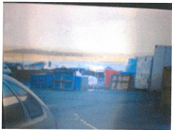A low-quality, blurry photograph showing a car in the foreground and blue and red structures or containers in the background against a bright sky.