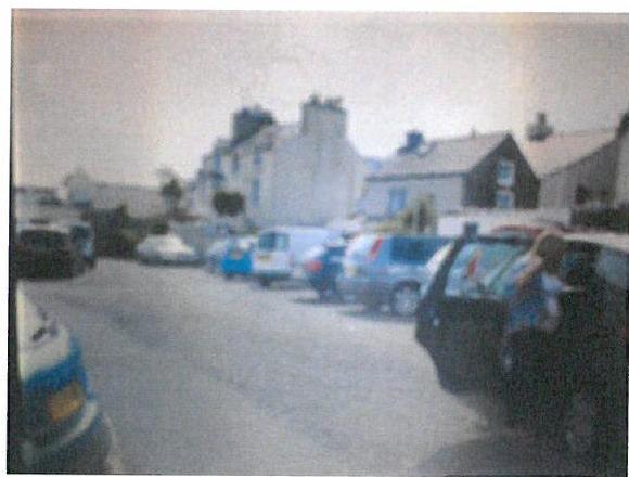 A low-resolution photograph showing a street scene with several parked cars and residential buildings in the background.