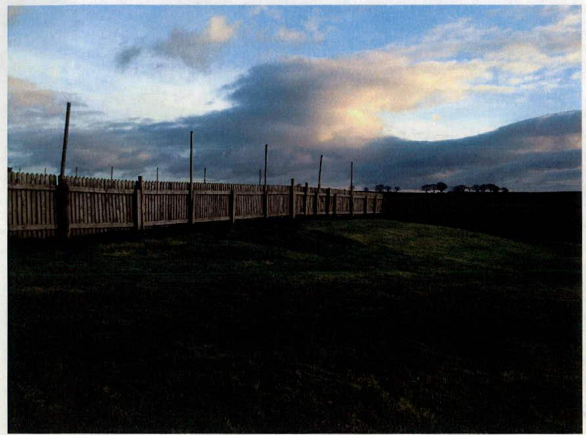 A photograph showing a wooden fence line running across a grassy field under a cloudy sky.