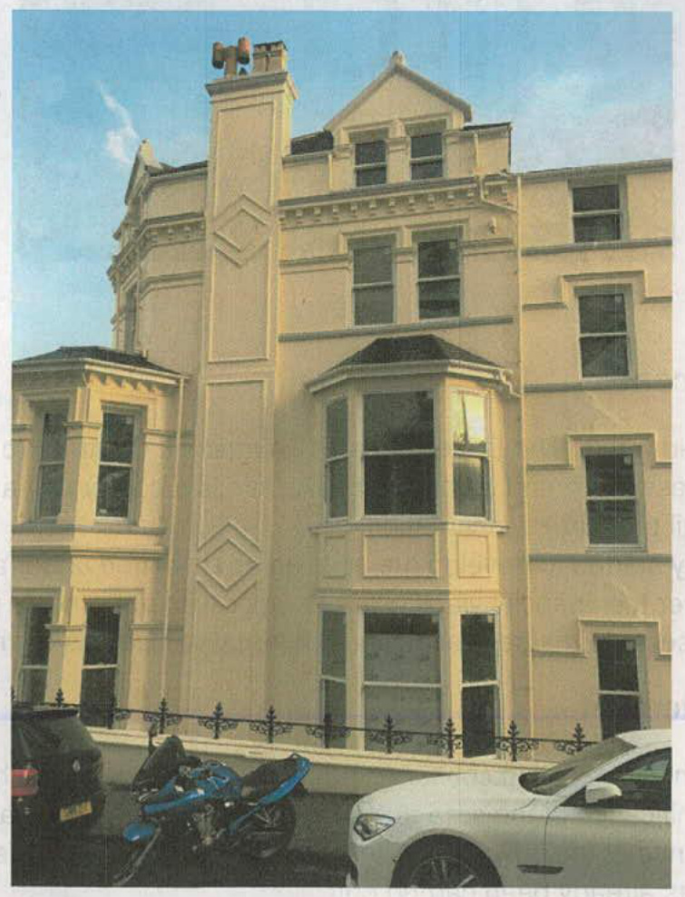 A street-level photograph showing the cream-colored facade of a large, multi-story residential building with bay windows and decorative stonework.