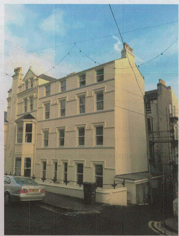A photograph showing the exterior facade of a multi-story cream-colored building, likely a block of flats or apartments, situated on a street with overhead wires.